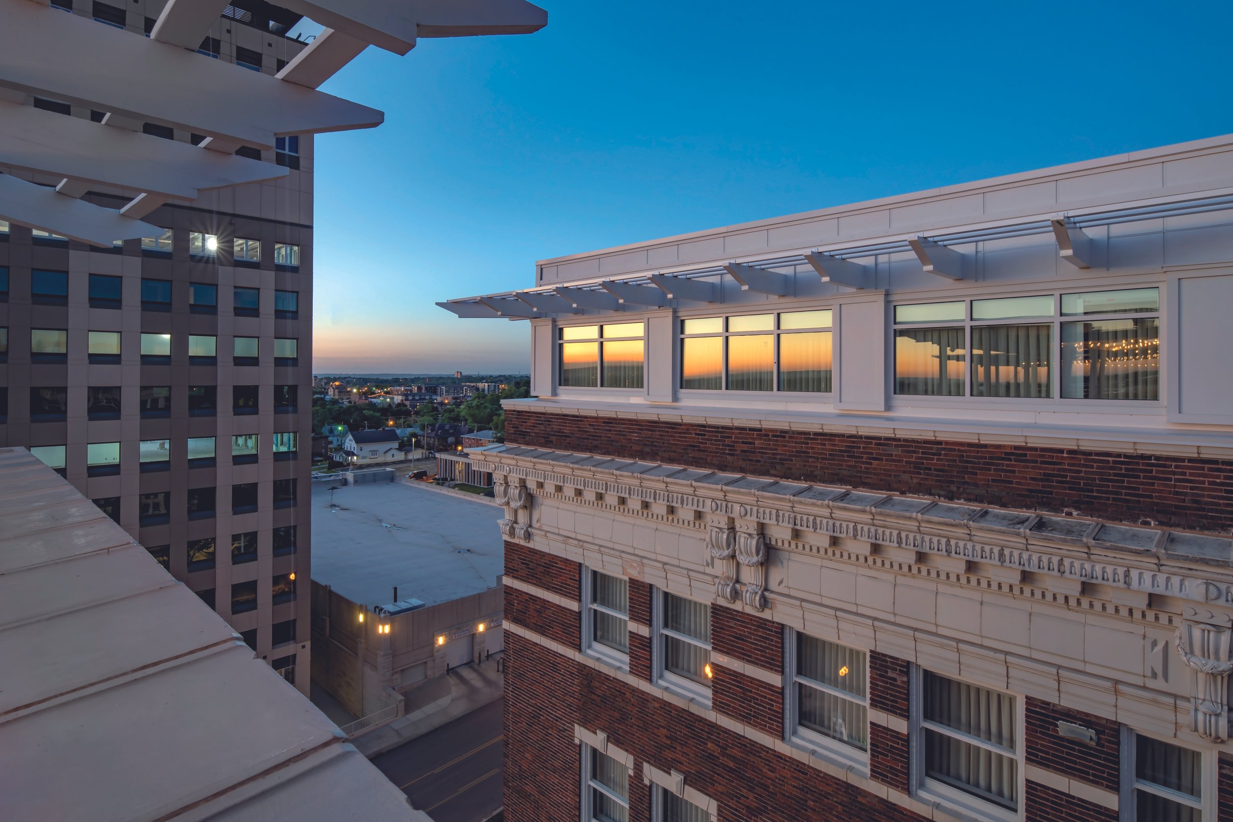 View from a rooftop at sunset showing neighboring buildings, including a brick building with large windows and decorative architectural elements, and a modern high-rise with numerous windows, under a clear blue sky with the sun setting on the horizon.