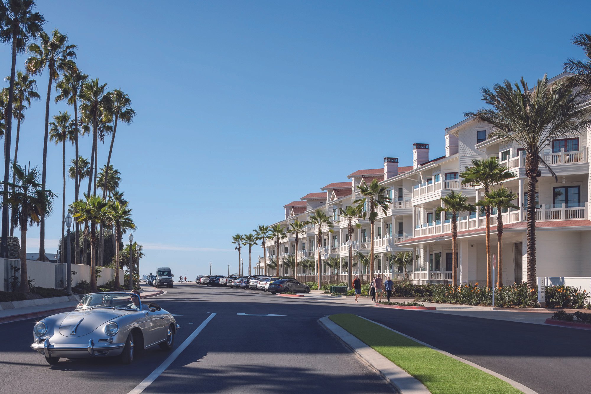 A street adjacent to the Hotel del Coronado lined with tall palm trees, modern white beachside apartments with balconies, parked cars, and a vintage convertible car in the foreground. Clear blue sky overhead.