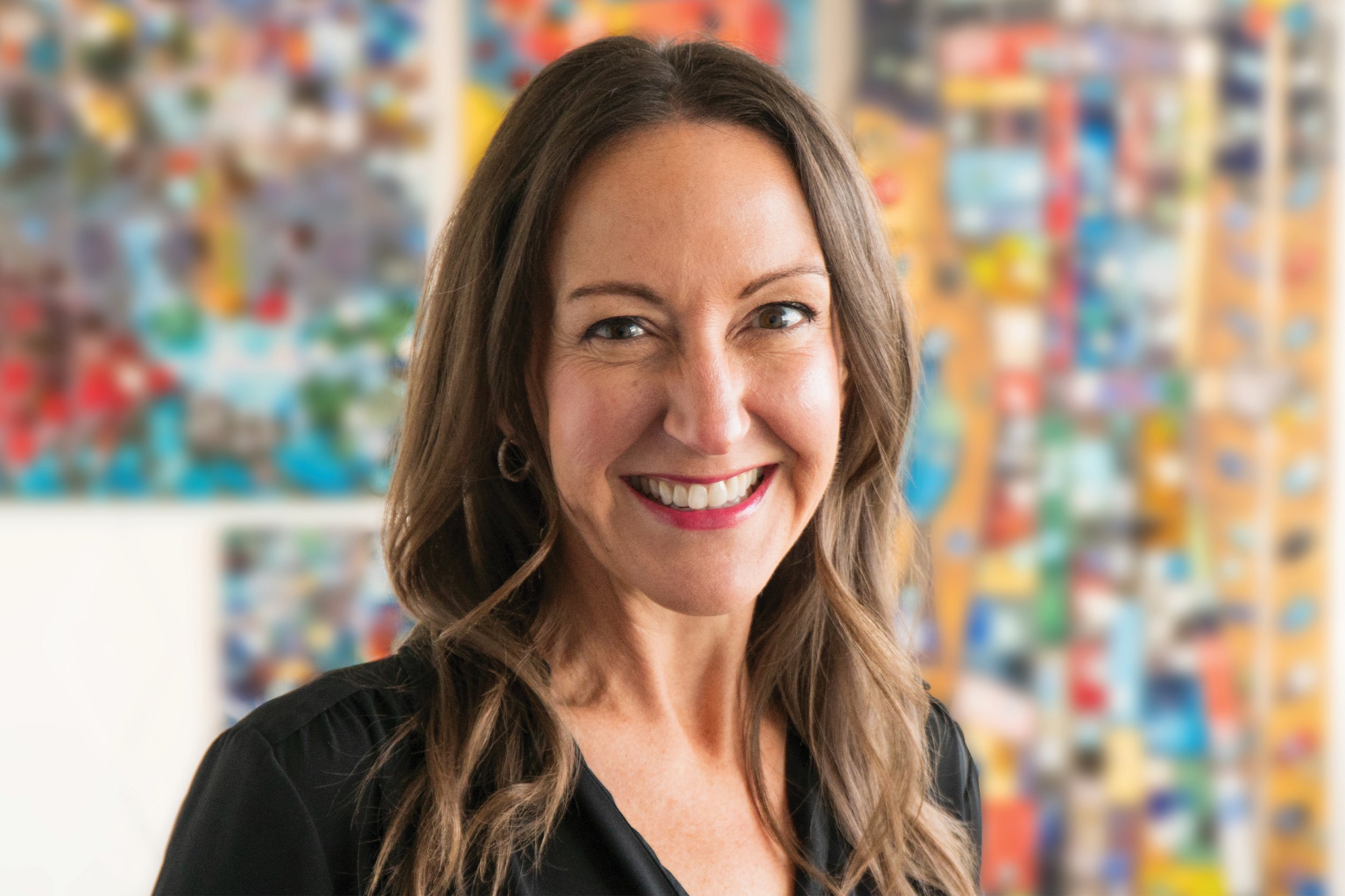 A woman with long brown hair smiling at the camera. She is wearing a black top and earrings. The background is colorful and blurred.