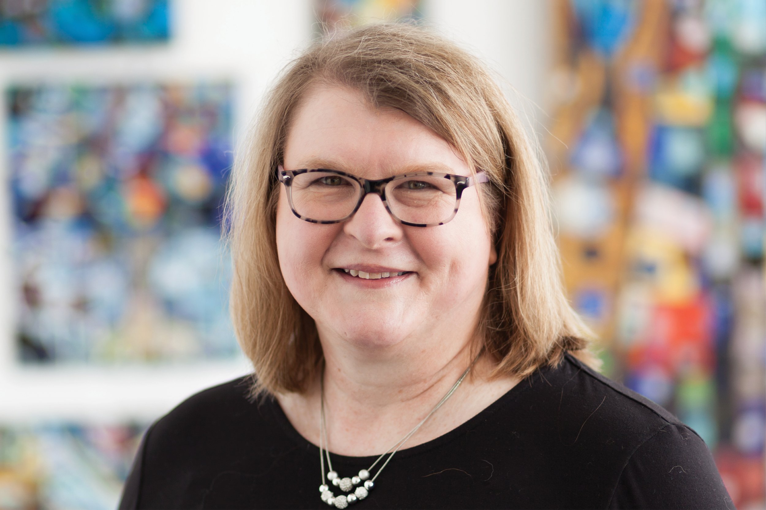 A smiling woman with glasses and shoulder-length light brown hair, wearing a black top and a pearl necklace, standing in front of colorful artwork.
