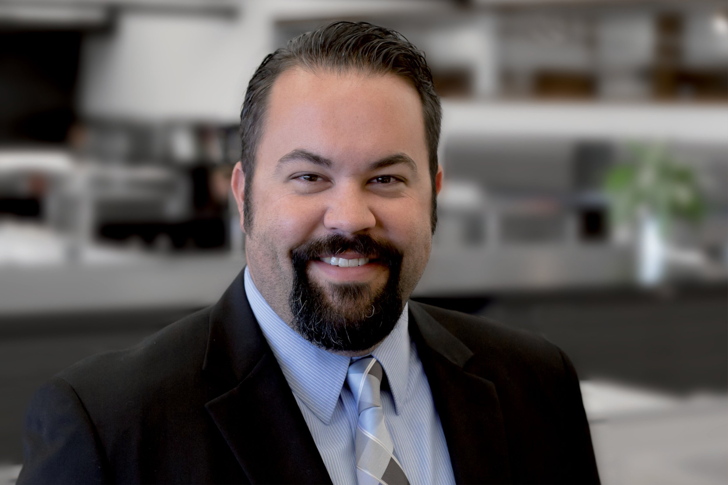 A smiling man with dark hair, a beard, and a mustache, wearing a black suit and a striped shirt and tie. He is indoors with blurred background of office or conference room.
