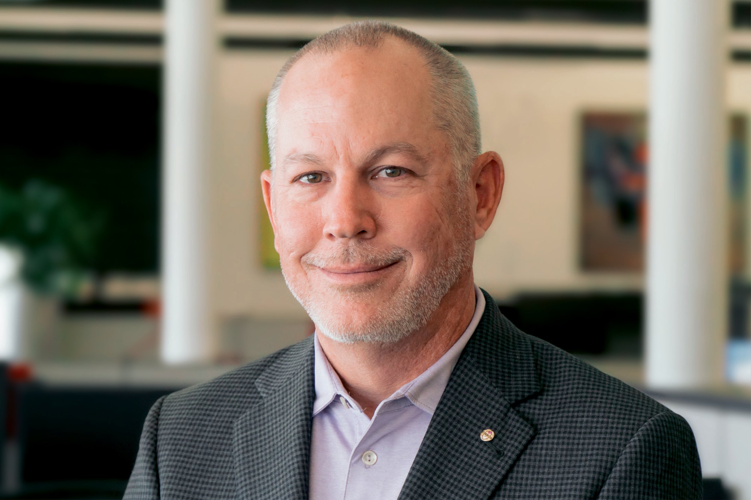 Portrait of Jay Brader, CFO, wearing a gray checkered blazer and a white collared shirt, in an indoor office setting.