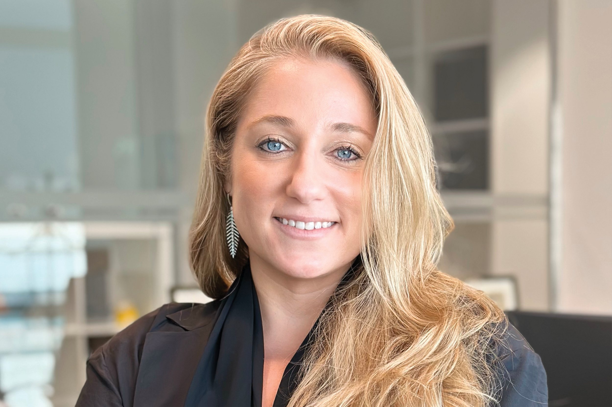 A woman with long blonde hair, blue eyes, and a bright smile in an office setting.