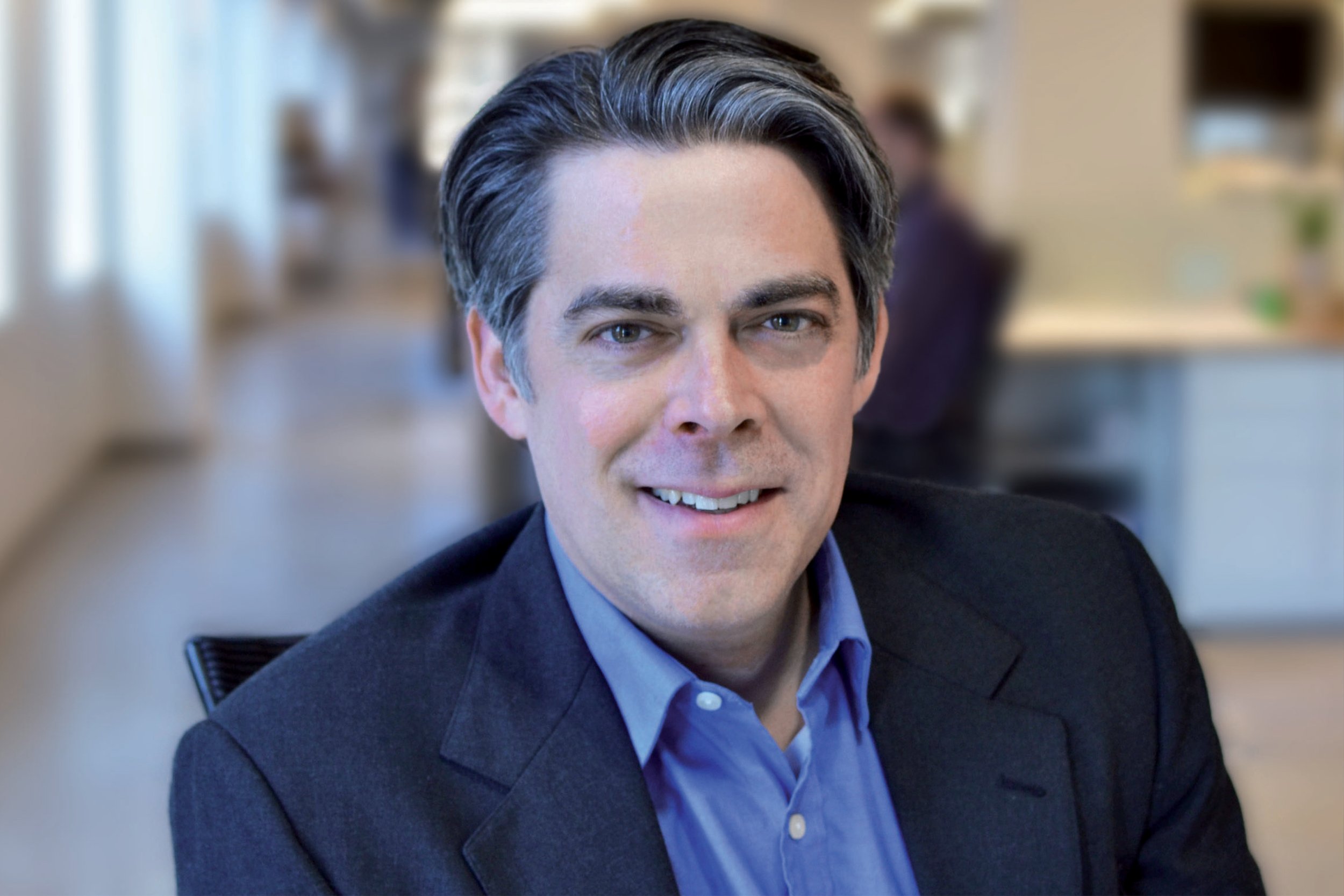 A professional man with dark and gray hair, wearing a dark blazer and a blue shirt, smiling at the camera in an office setting with blurred background.