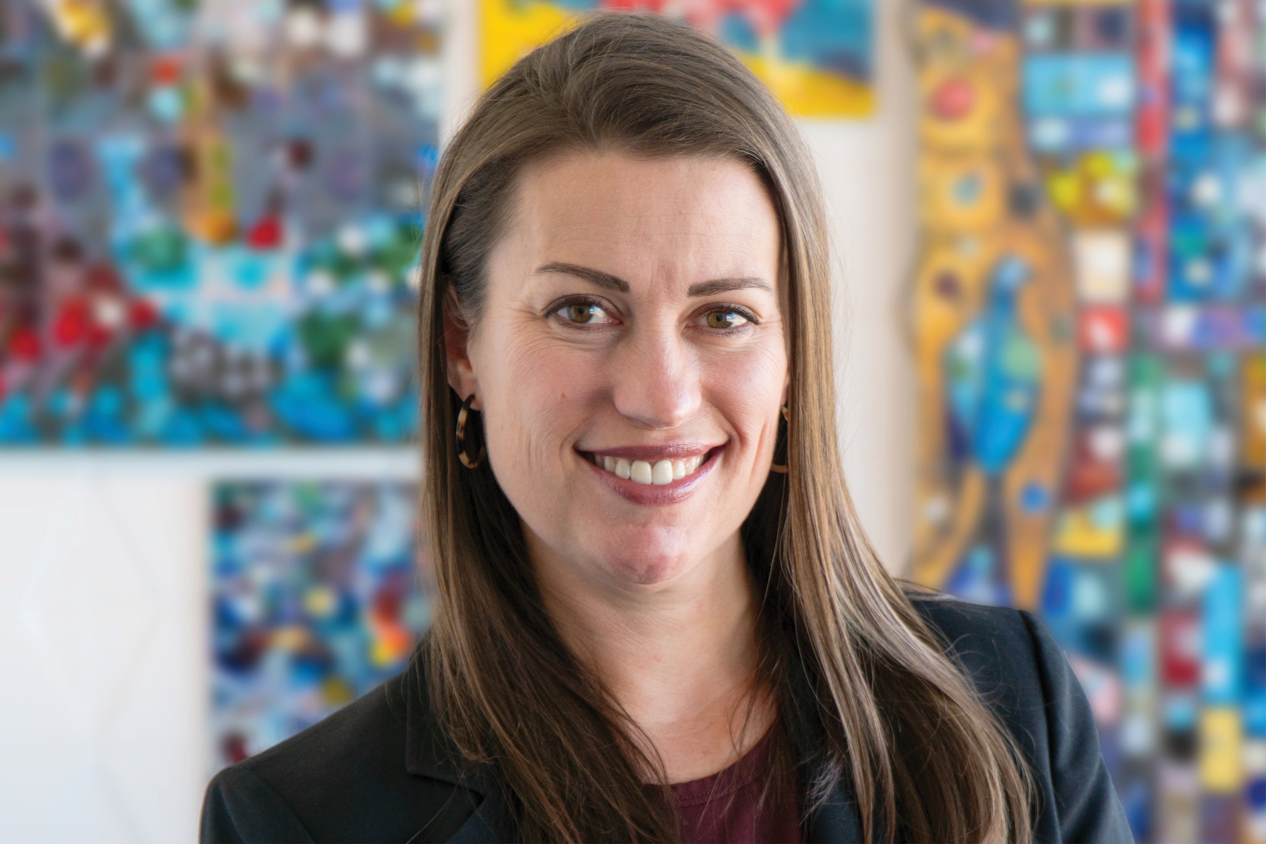 A woman with brown hair and earrings smiling in front of a colorful abstract background.