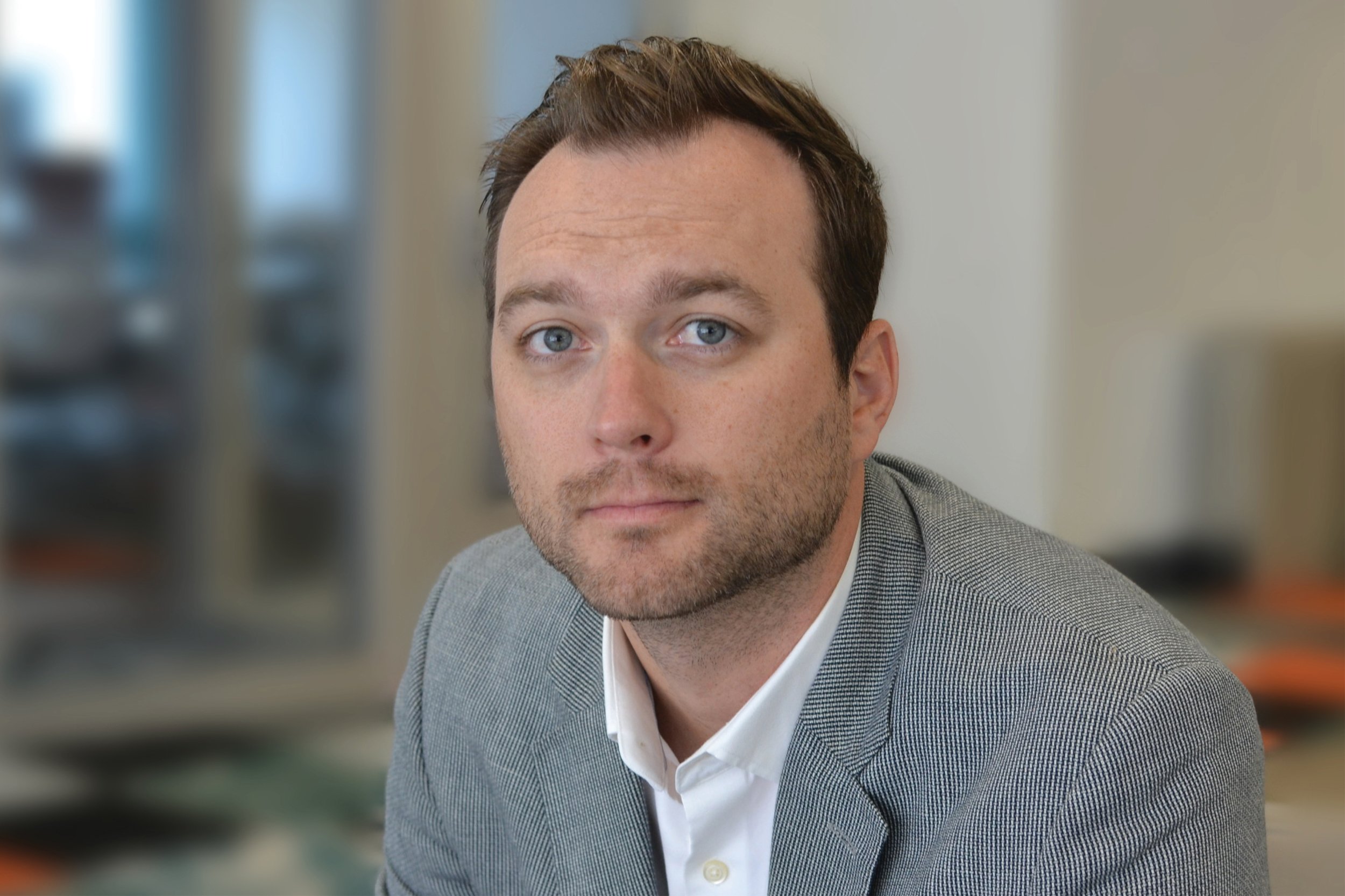 Portrait Linn Bjornrud wearing a gray blazer and white collared shirt, seated indoors with blurred background.