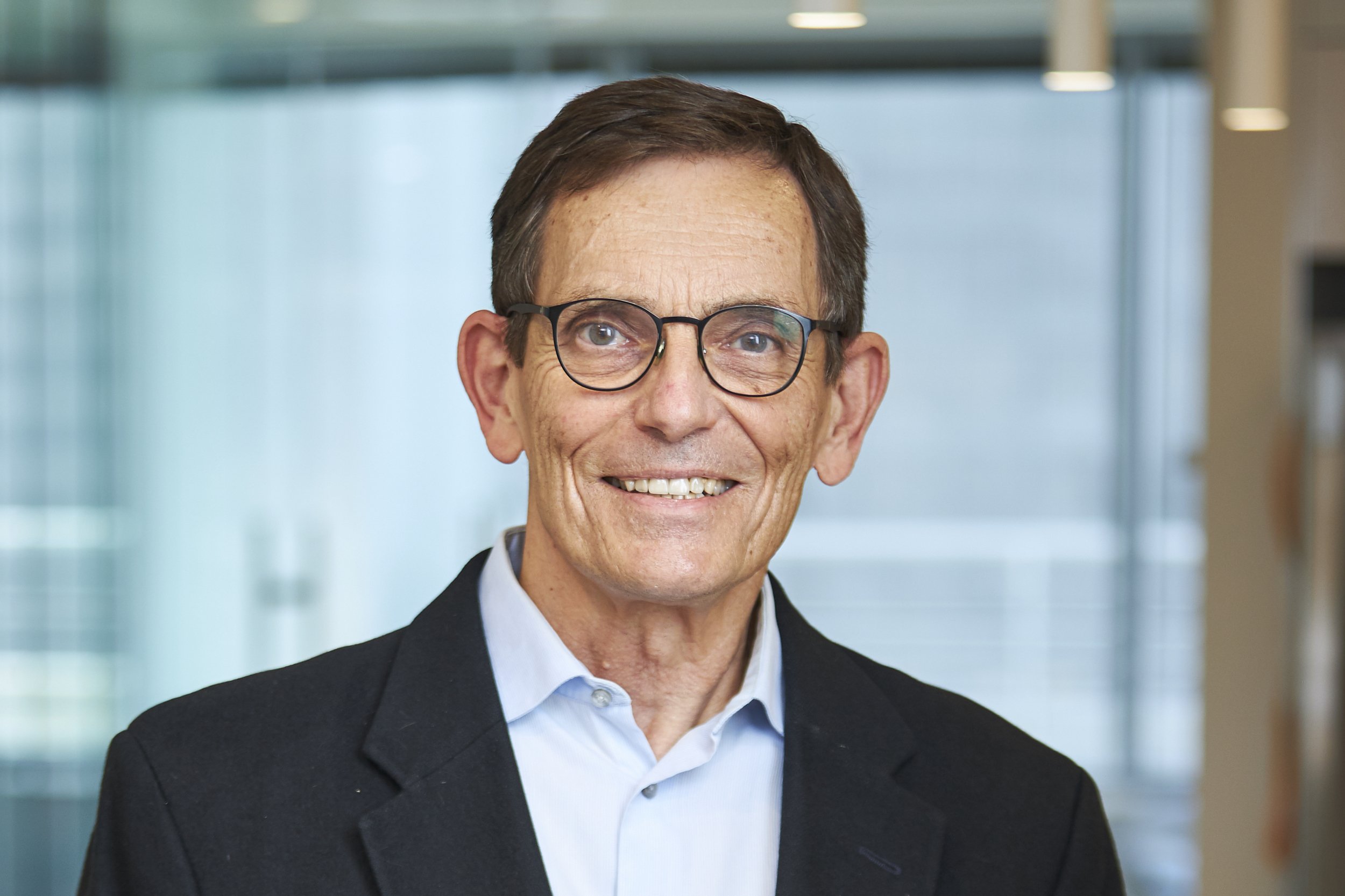 Headshot of a middle-aged man with glasses, smiling, wearing a suit jacket and a light blue shirt, office background with glass windows.