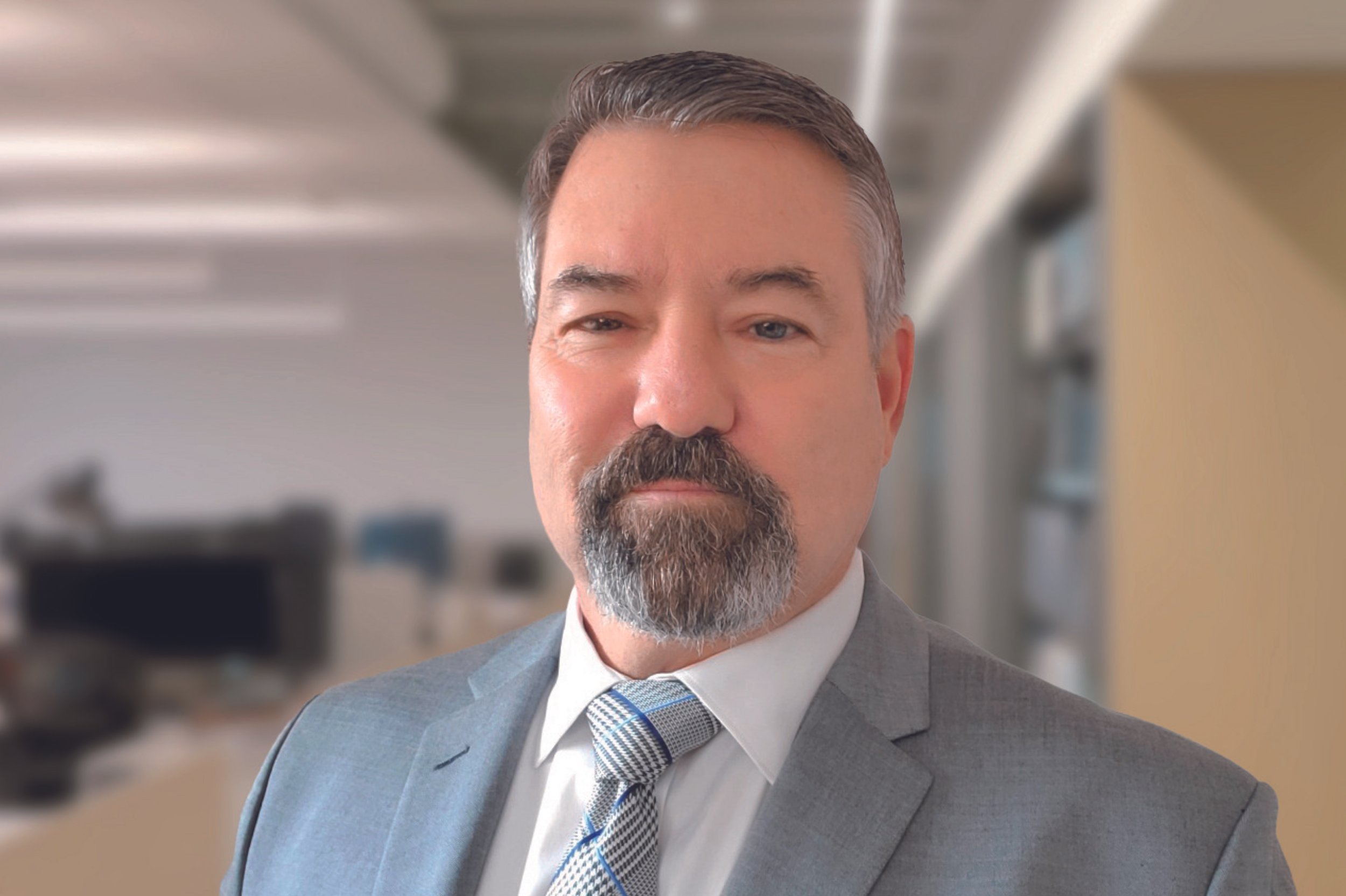 A middle-aged man with a beard and short hair, wearing a gray suit and a patterned tie, in an office environment.