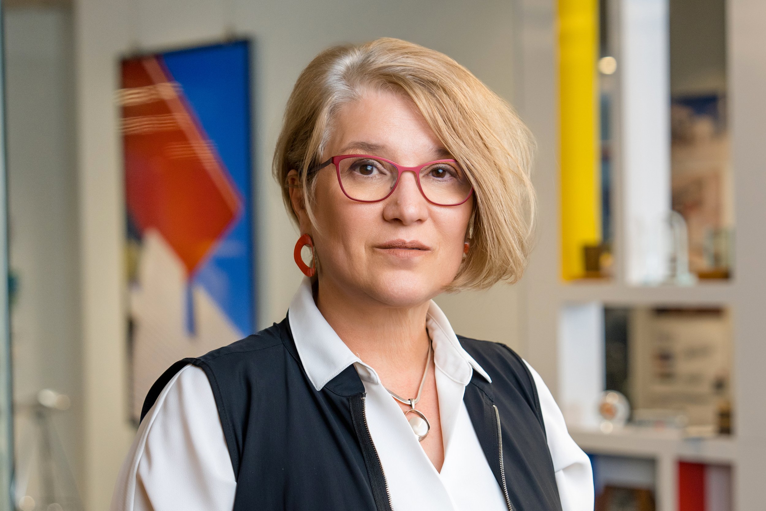 Portrait of Irena Savakova, Design Principal, wearing red glasses, a white shirt, and a black vest, standing indoors with colorful abstract art and a yellow lamp in the background.